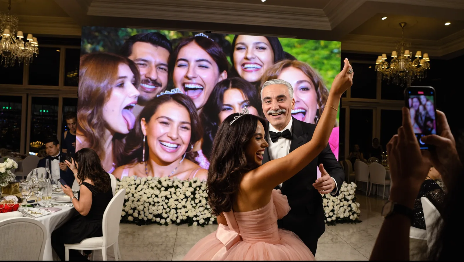 Salón de noche: la quinceañera baila con su papá frente a la pantalla del salón, donde aparece la selfie que acaban de subir sus amigas