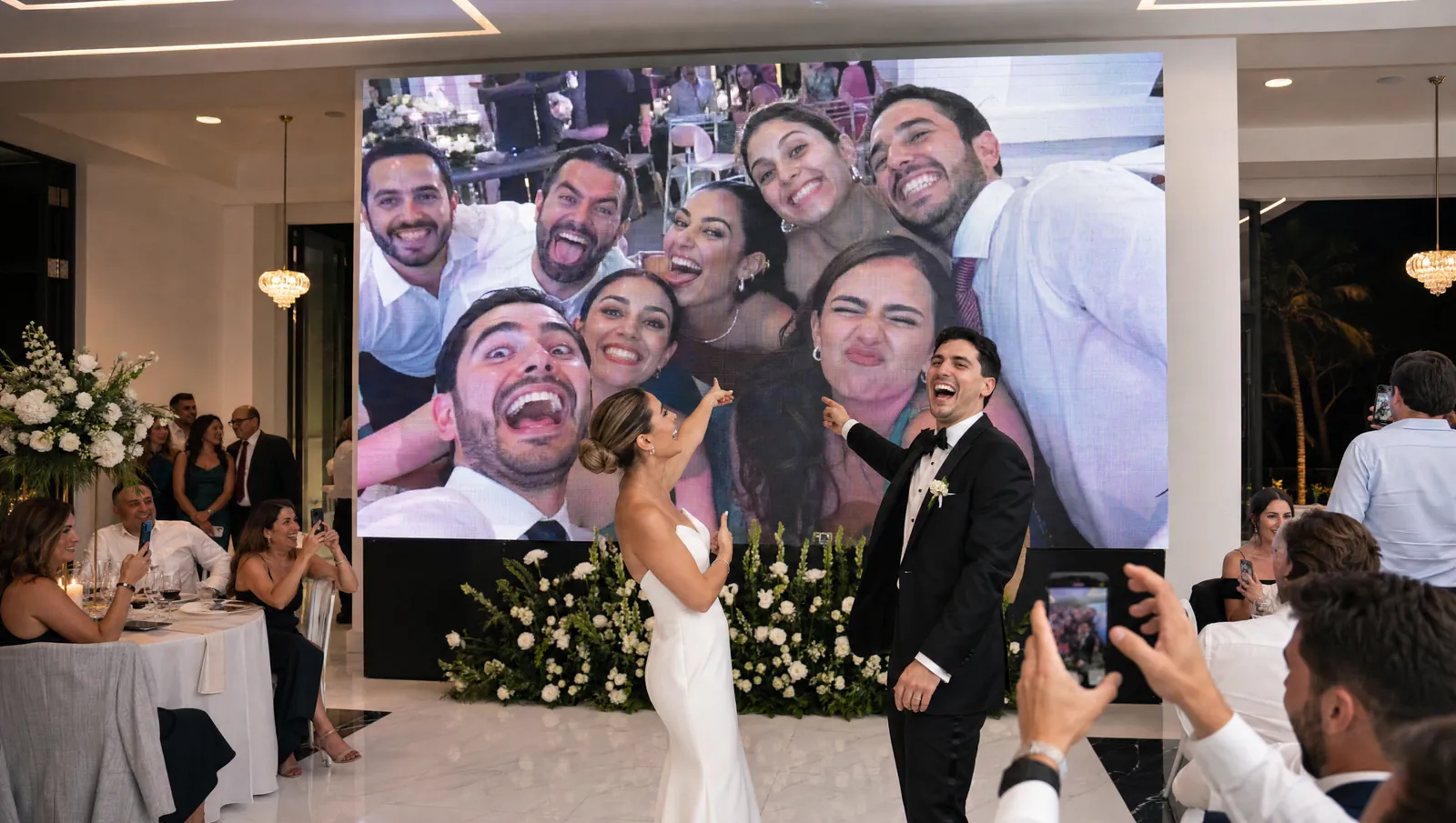Salón moderno de noche: los novios se ríen y señalan la pantalla gigante, donde aparece la selfie que acaban de sacar sus invitados en la mesa
